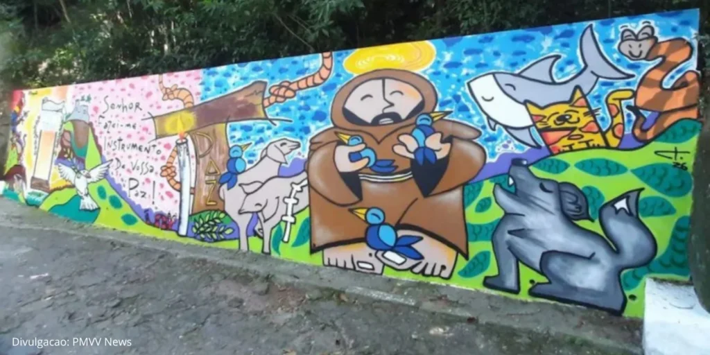 Mural colorido pintado para a Festa da Penha 2026 com ilustrações religiosas e elementos da natureza, incluindo São Francisco, animais e símbolos de paz, em parede externa cercada por vegetação.