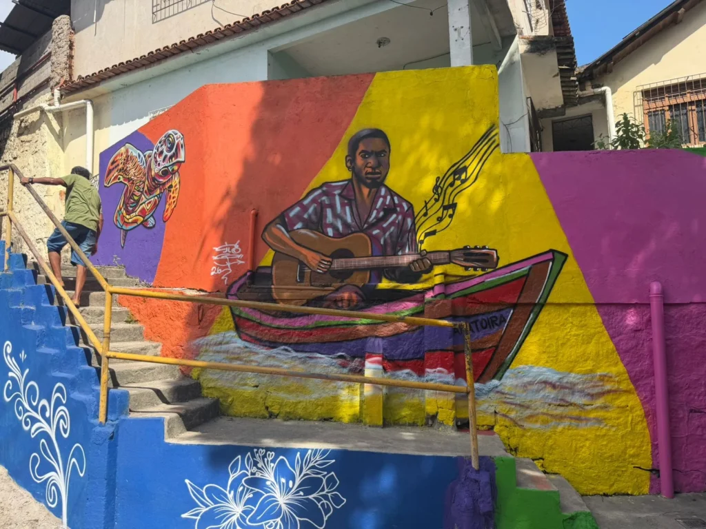 Grafite urbano com músico tocando violão em barco e tartaruga marinha ao lado, em mural colorido do projeto Vizinho da Arte, realizado em Caratoíra, Vitória-ES, com apoio da Politintas.