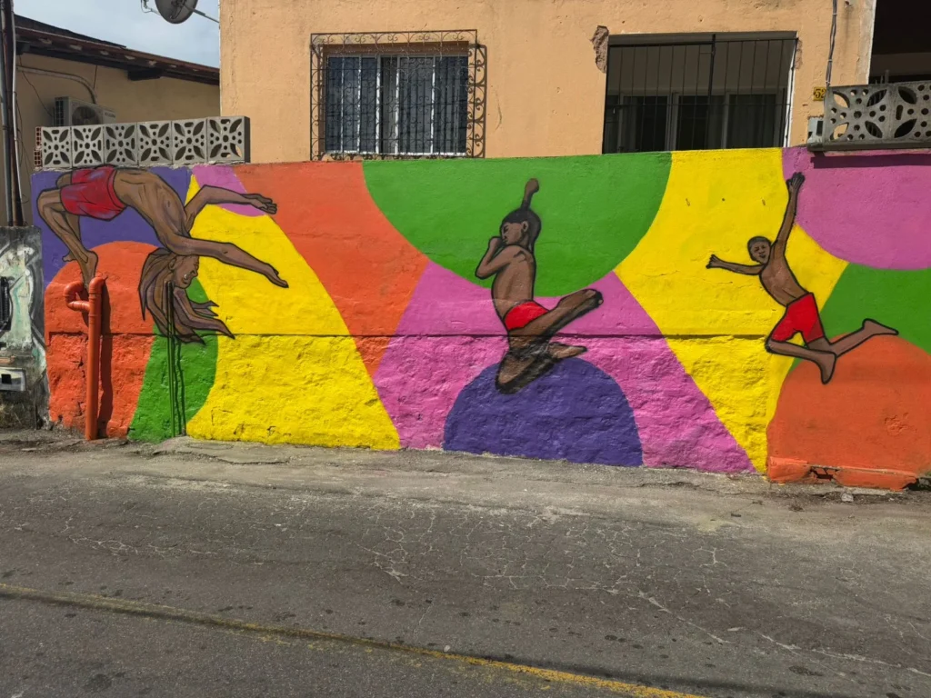  Mural de grafite com crianças saltando e brincando sobre fundo geométrico colorido, retratando lazer e infância na comunidade de Caratoíra, em Vitória-ES, no projeto Vizinho da Arte apoiado pela Politintas.