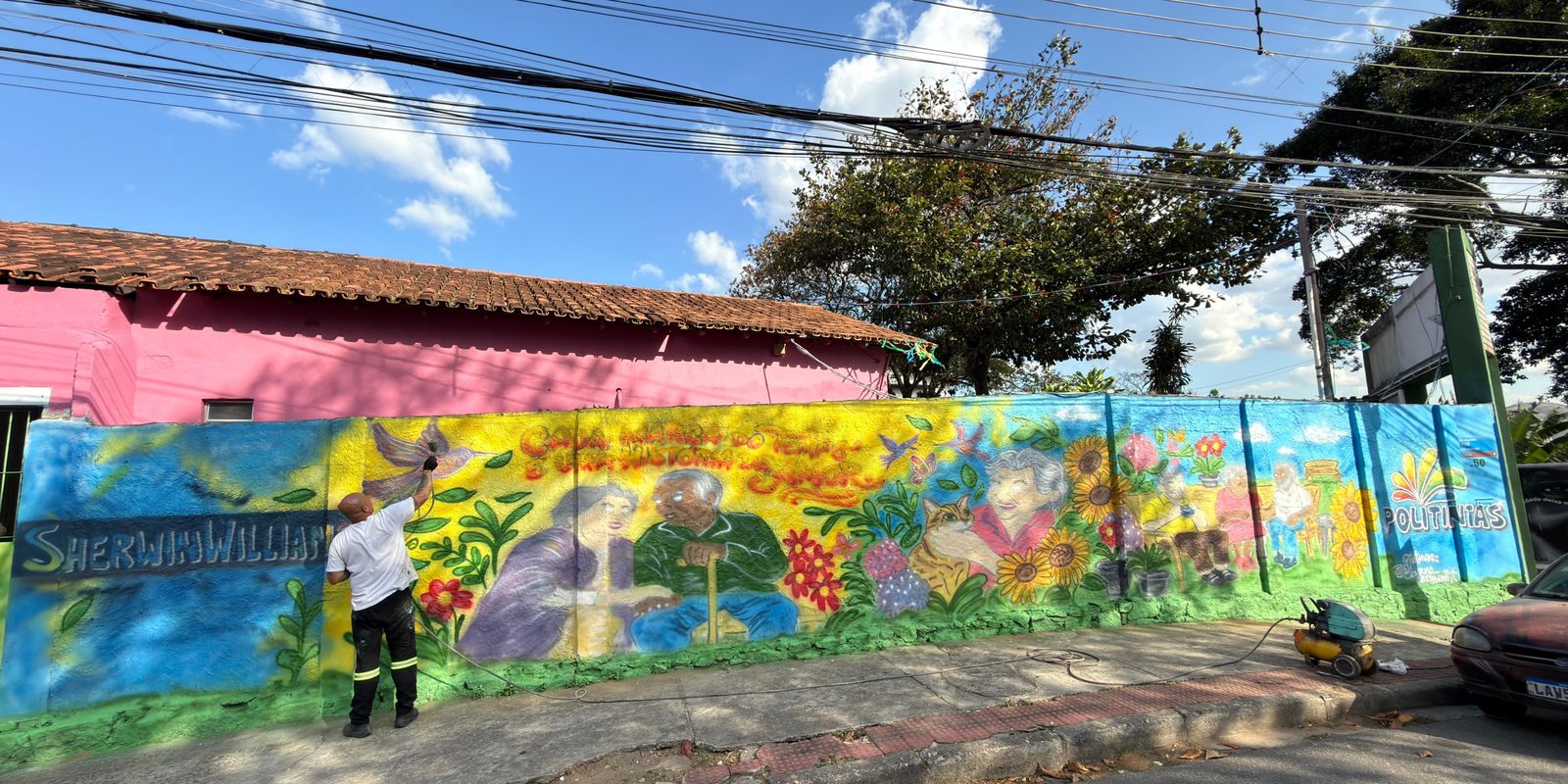 Artista pinta mural colorido no Asilo dos Idosos de Vitória, retratando idosos, flores e natureza durante ação voluntária de revitalização dos espaços apoiada pela Politintas.