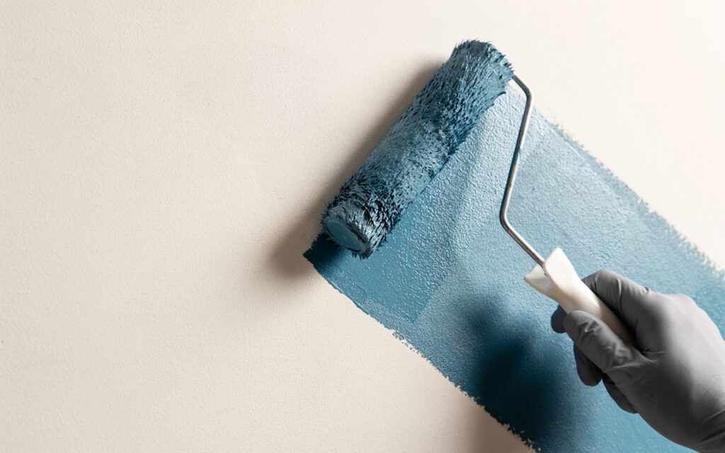 Mão com luva usando rolo para aplicar tinta azul em parede clara, mostrando processo de pintura e renovação de ambientes internos.